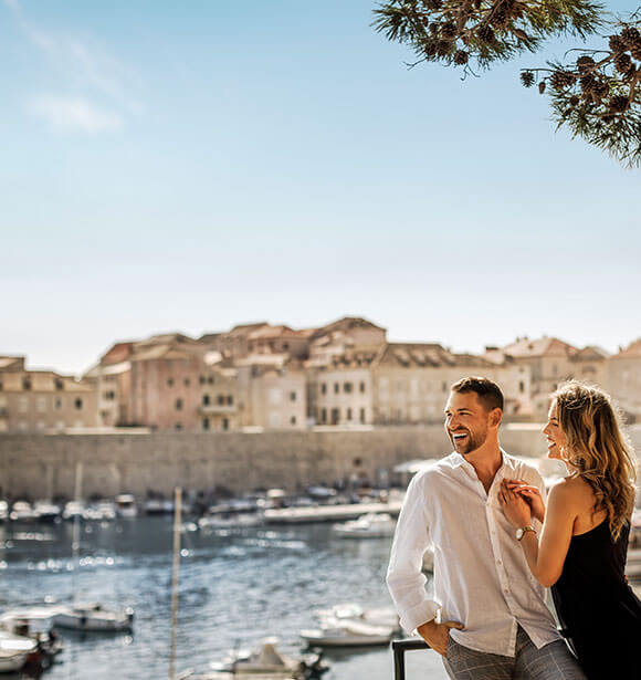 Ein Mann und eine Frau, die am Meer stehen, und im Hintergrund Boote und Gebäude
